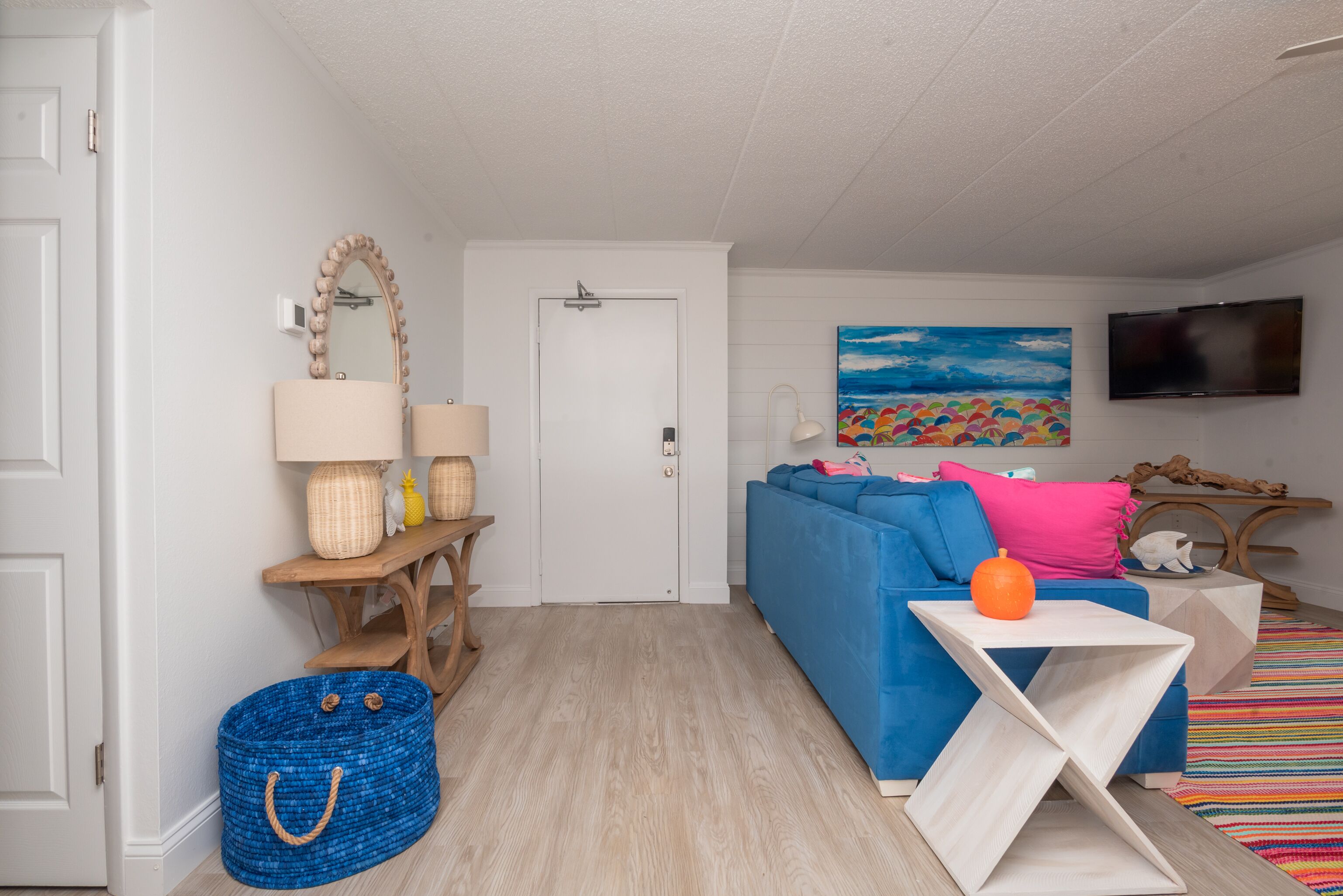 Een moderne woonkamer met een blauwe bank, een witte bijzettafel, een houten bankje, een blauwe geweven mand en een kleurrijk schilderij aan de muur.