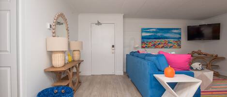 Een moderne woonkamer met een blauwe bank, een witte bijzettafel, een houten bankje, een blauwe geweven mand en een kleurrijk schilderij aan de muur.