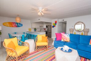 A bright living room with a blue sofa, yellow and blue chairs, a colorful striped rug, and a kitchen in the background.