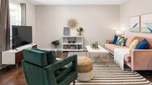 Ein modernes Wohnzimmer mit einem Flachbildfernseher, einem grünen Sessel, einem rosa Sofa mit bunten Kissen, einem Streifen Teppich und einem weißen Konsolentisch.