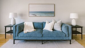 A blue sofa with a white throw blanket, flanked by two black nightstands with white lamps, against a plain wall with a framed landscape painting.