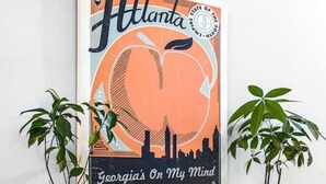 Une grande peinture murale représentant Atlanta, encadrée, avec une vue sur les gratte-ciel de la ville et un cœur, placée au-dessus d’une table en bois sur laquelle reposent deux plantes en pot.