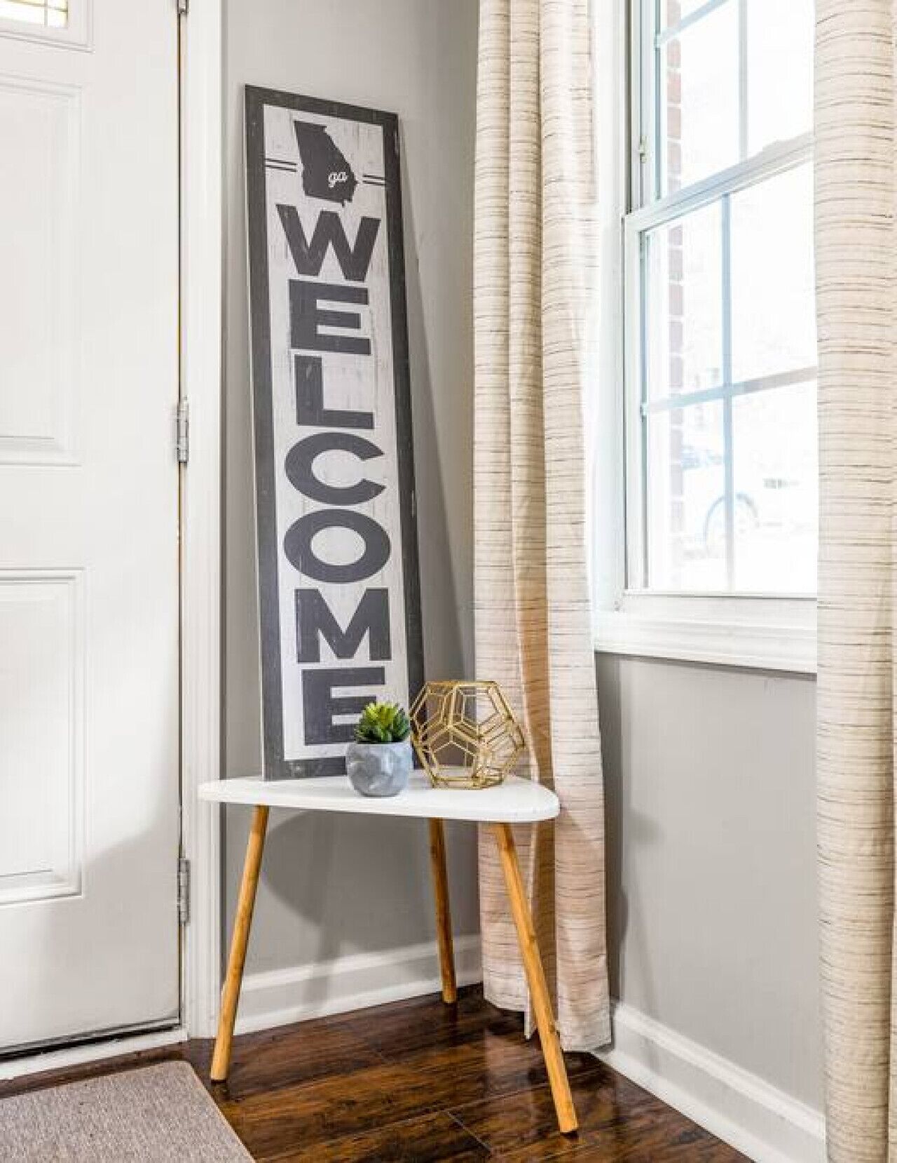 A vertical welcome sign on a white stand with a small potted plant and decorative item beside it, next to a window with curtains.