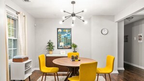 Un coin repas avec une table ronde en bois, quatre chaises jaunes, un lustre moderne, une affiche représentant un paysage urbain et une horloge au mur.