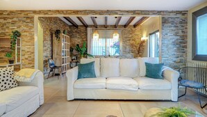 A spacious living room with a white sofa, a smaller white armchair, and a patterned armchair. The room features exposed wooden beams, stone walls, and large windows allowing natural light. - Villa Nirvana by Hello Homes Sitges (Sitges)