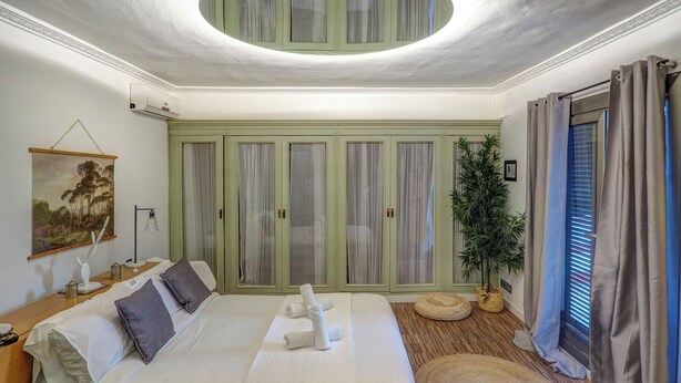 A bedroom with a large bed, a bedside table, a lamp, a painting, a potted plant, and a window with curtains. - Villa Nirvana by Hello Homes Sitges (Sitges)