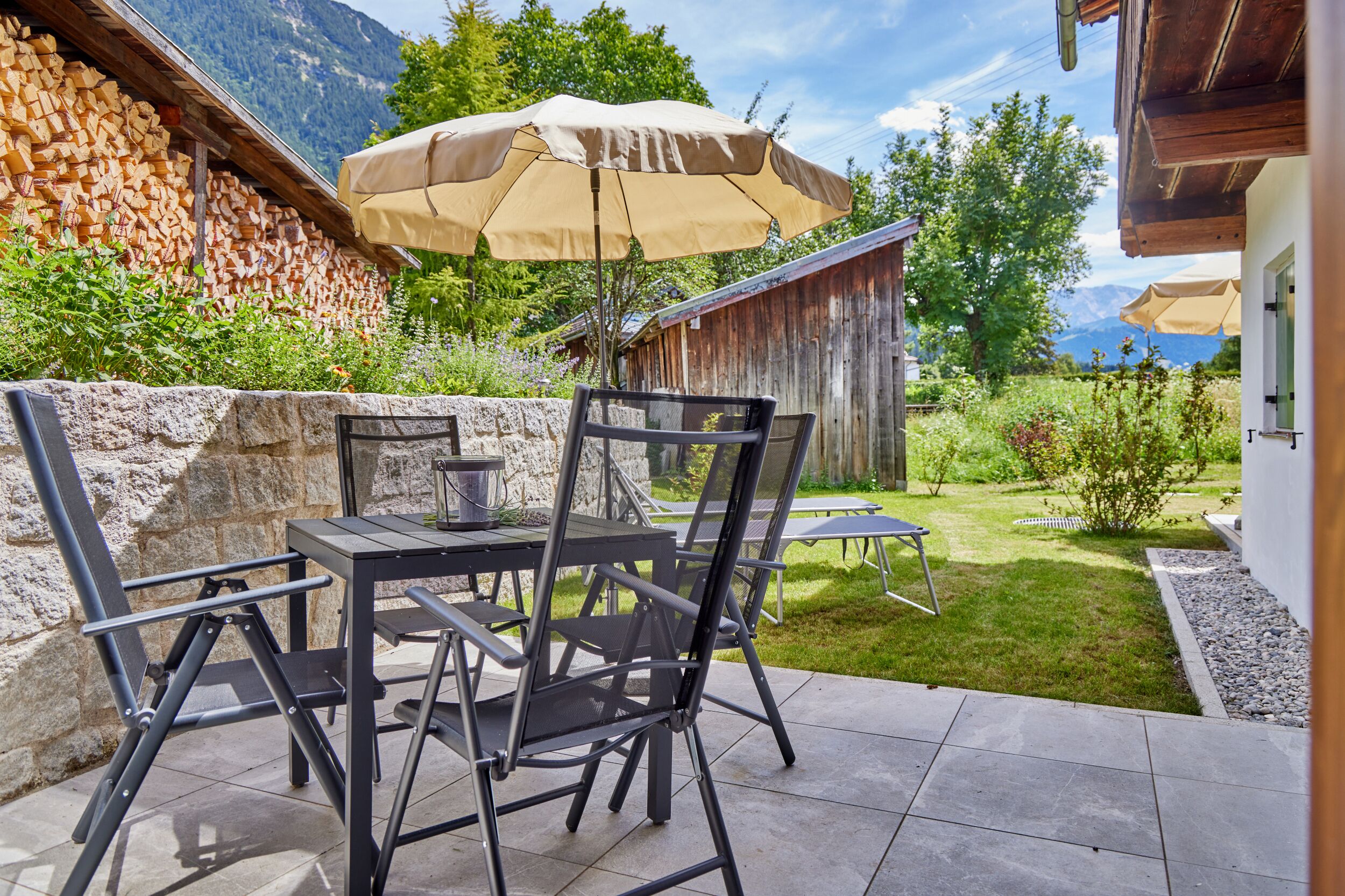 Une terrasse avec une table et des chaises, un parasol et un mur de pierres sur lequel est empilé du bois de chauffage.