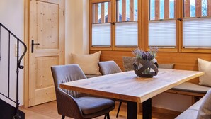 Uma sala de jantar com mesa de madeira, cadeiras e um lustre. Há janelas amplas com persianas, uma porta e uma escada.