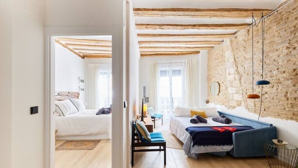 Une chambre moderne avec un grand lit, un petit canapé et un bureau. La pièce est dotée de poutres apparentes, d’un mur en briques et de grandes fenêtres.