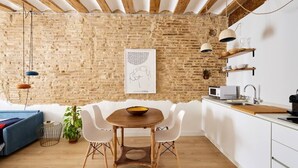 Une cuisine moderne avec un mur en briques, des poutres en bois au plafond, des meubles blancs et un coin repas comprenant une table en bois et des chaises blanches.