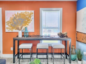 Un coin repas comprenant une table, trois chaises, une fenêtre avec des stores et un grand tableau abstrait accroché au mur.