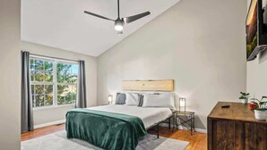 A bedroom with a bed, a desk, a ceiling fan, a window, and a view of trees.