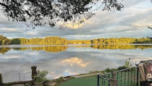 A serene lakeside view with a wooden deck, a green lawn, and a nest on a tree.