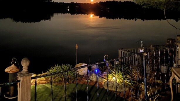 A nighttime view of a lake with a sunset, a dock, and a well-lit garden area with plants and a small pond.