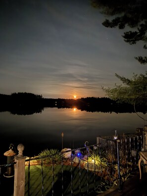Ein Nachtblick auf einen See mit Sonnenuntergang, einem Steg und einem gut beleuchteten Gartenbereich mit Pflanzen und einem kleinen Teich.