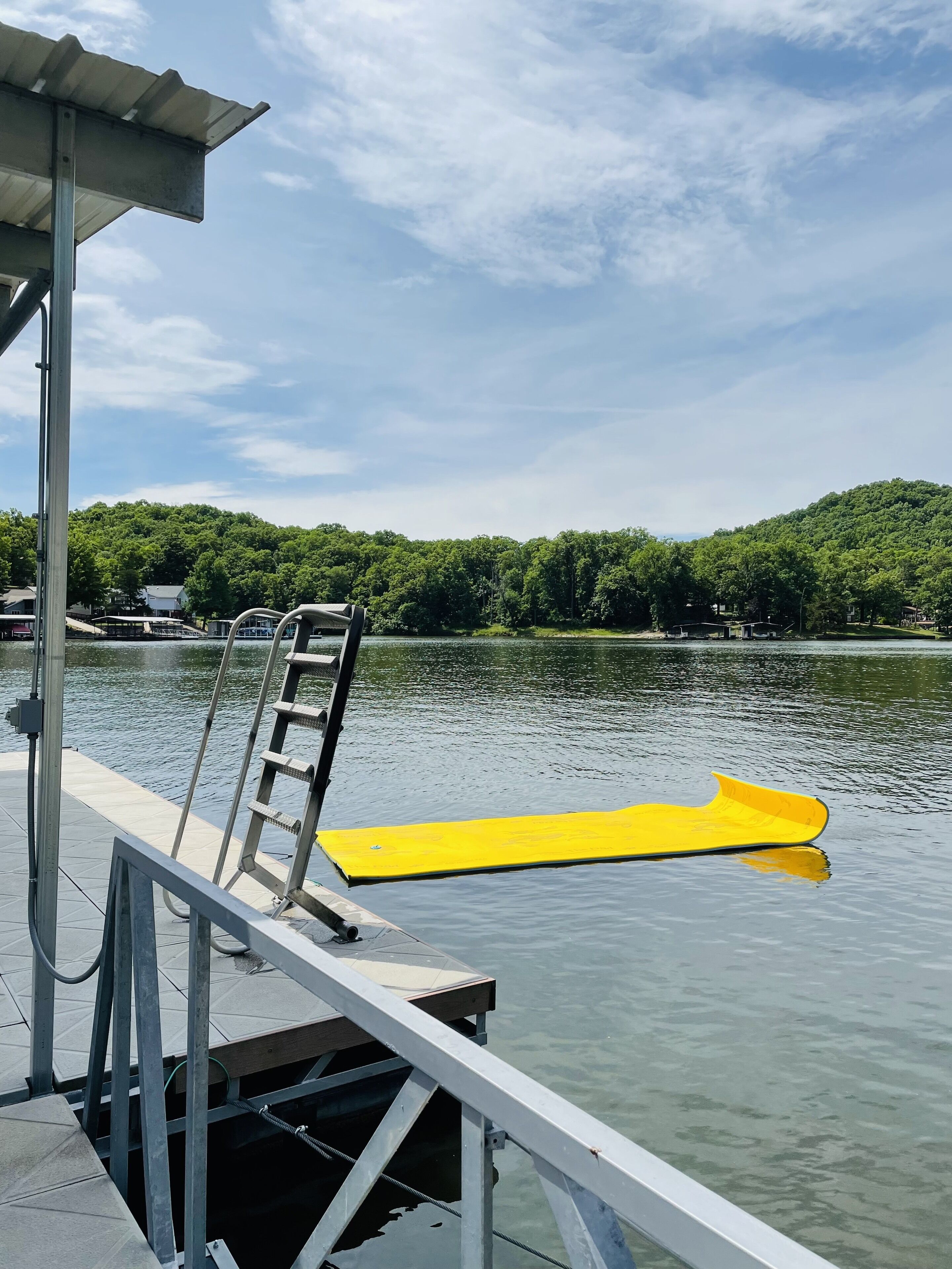 Een steiger met een geel paddleboard, een metalen ladder en een overdekt zitgedeelte met uitzicht op een meer met bomen.