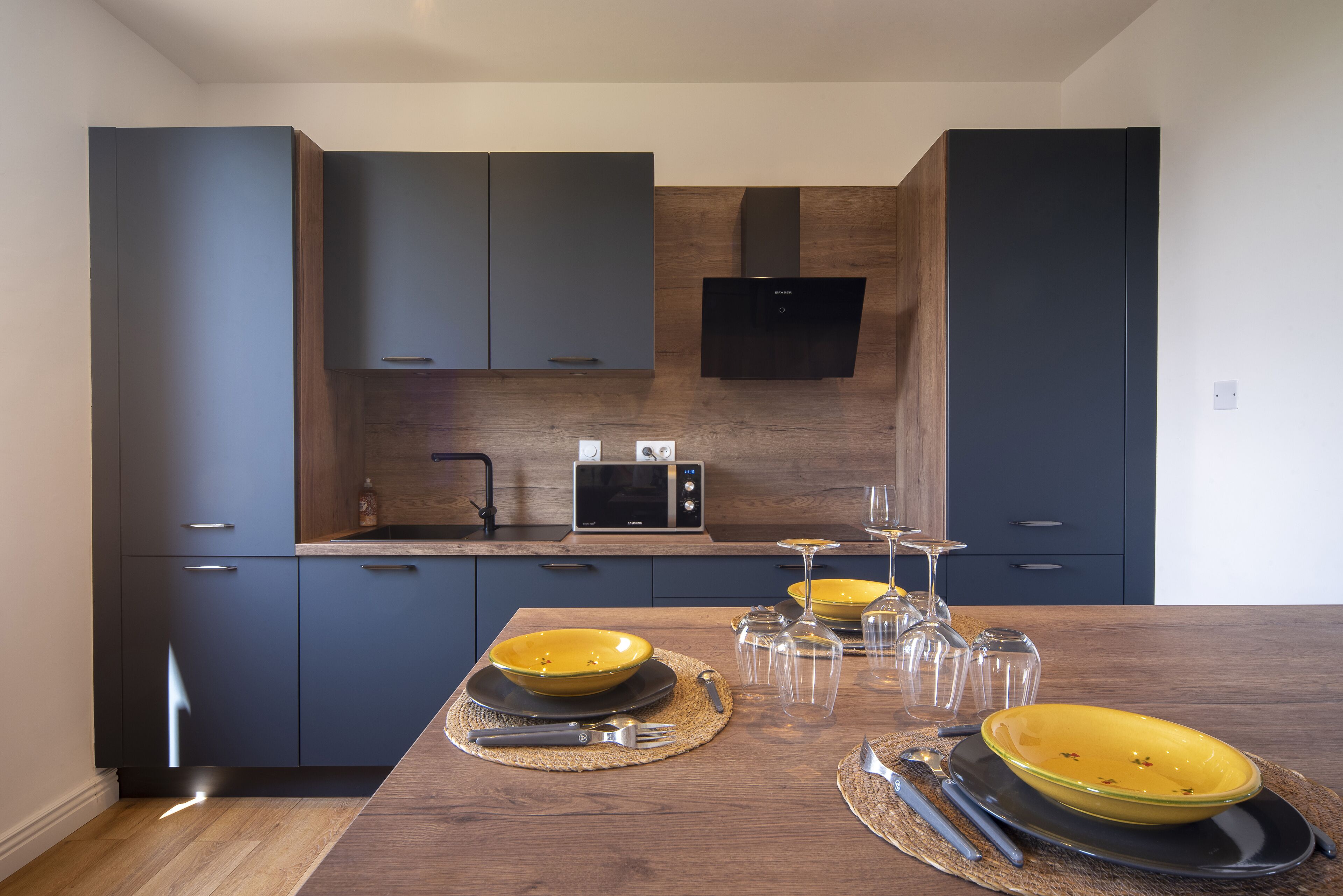 Une cuisine moderne avec des meubles de couleur sombre, un four à micro-ondes et une table à manger agrémentée de bols et de couverts jaunes.