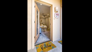Un couloir avec un paillasson sur lequel est écrit « Bonjour », un ventilateur de plafond, un lit et un panneau mural portant le numéro 102.