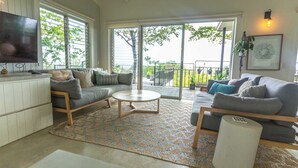 A spacious living room with a large sofa, a coffee table, and a TV. There is a sliding glass door leading to a balcony with a view of greenery. - Casa Aves - Gorgeous Ocean & Jungle View 3BR w/ Pool (nosara)