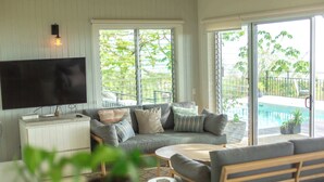 A living room with a grey sofa, a flat-screen TV, and a pool view. - Casa Aves - Gorgeous Ocean & Jungle View 3BR w/ Pool (nosara)