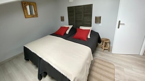 Un dormitorio con cama, dos almohadas rojas, una colcha blanca, un taburete de madera, una puerta y una lámpara colgante.