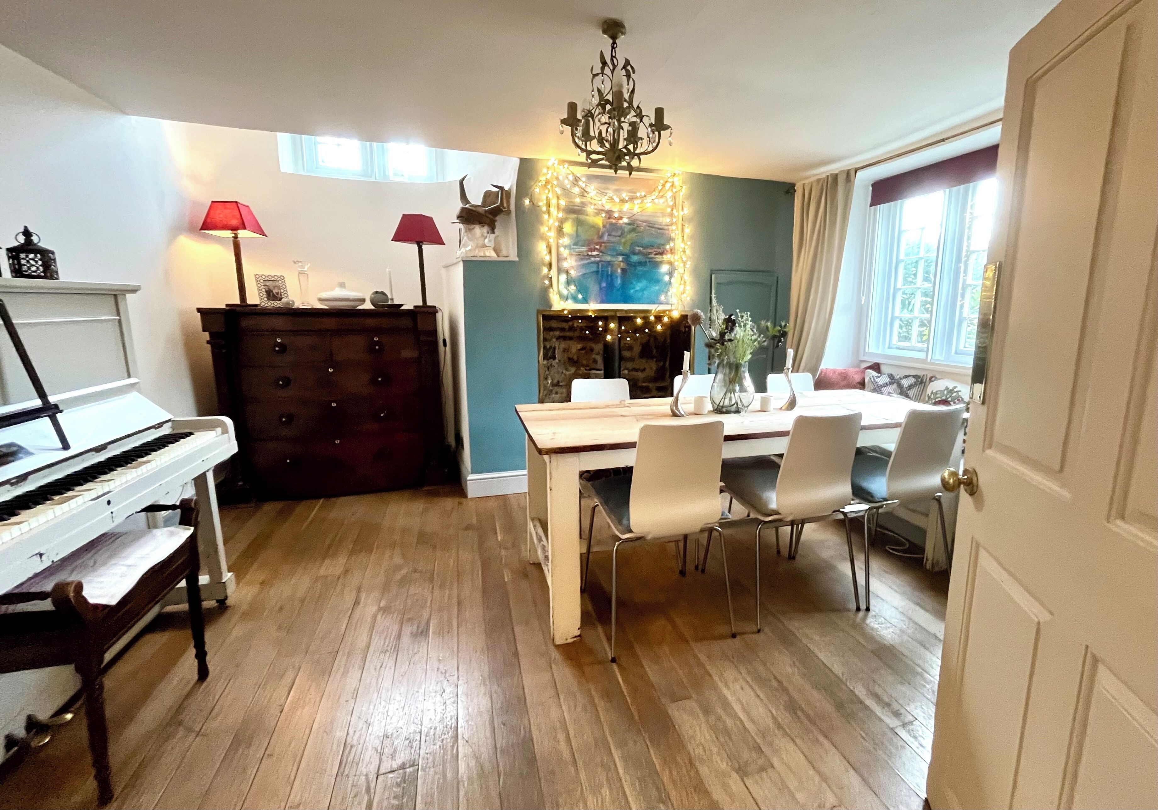 Un comedor con una mesa de madera, sillas blancas, una lámpara de araña y un piano.