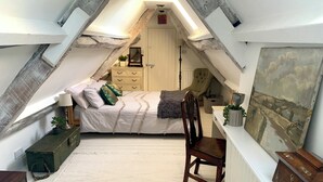 A cozy attic bedroom with a sloped ceiling, a bed with white bedding, a wooden chair, a dresser, and a painting on the wall.