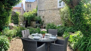 A round table with four wicker chairs, surrounded by a garden with various plants and a stone building in the background.