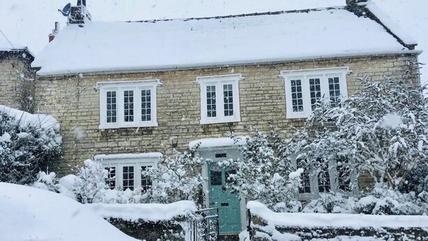A stone building with multiple windows and a green door, surrounded by snow-covered trees and a stone wall.