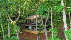 A treehouse with a wooden deck and hanging lights, surrounded by dense forest. - Treetop Getaway with AMAZING lake views! (Jasper)