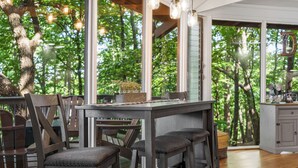 A wooden dining area with a high table and chairs, large windows with a view of trees, and a kitchen area with cabinets. - Treetop Getaway with AMAZING lake views! (Jasper)