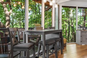 A wooden dining area with a high table and chairs, large windows with a view of trees, and a kitchen area with cabinets.