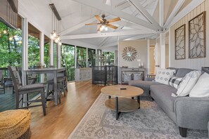 A spacious living area with a large sofa, a round coffee table, and a dining table with chairs. There is a ceiling fan, a decorative wall piece, and a view of greenery outside.