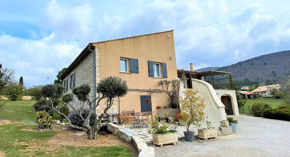A two-story house with a stone facade, a small patio with outdoor furniture, and a garden with various plants.