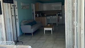 Uma sala de estar ampla, com um sofá, uma mesa de centro e uma mesa de jantar com toalha estampada. Uma cozinha com armários brancos e bancada é visível ao fundo.