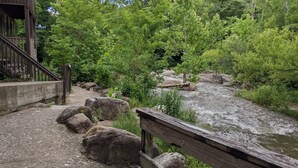 A wooden bridge over a river with a concrete walkway on the left, surrounded by lush greenery.