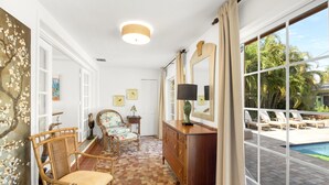 Een kamer met een zwembad, rieten stoelen, een houten dressoir en een groot raam met gordijnen.