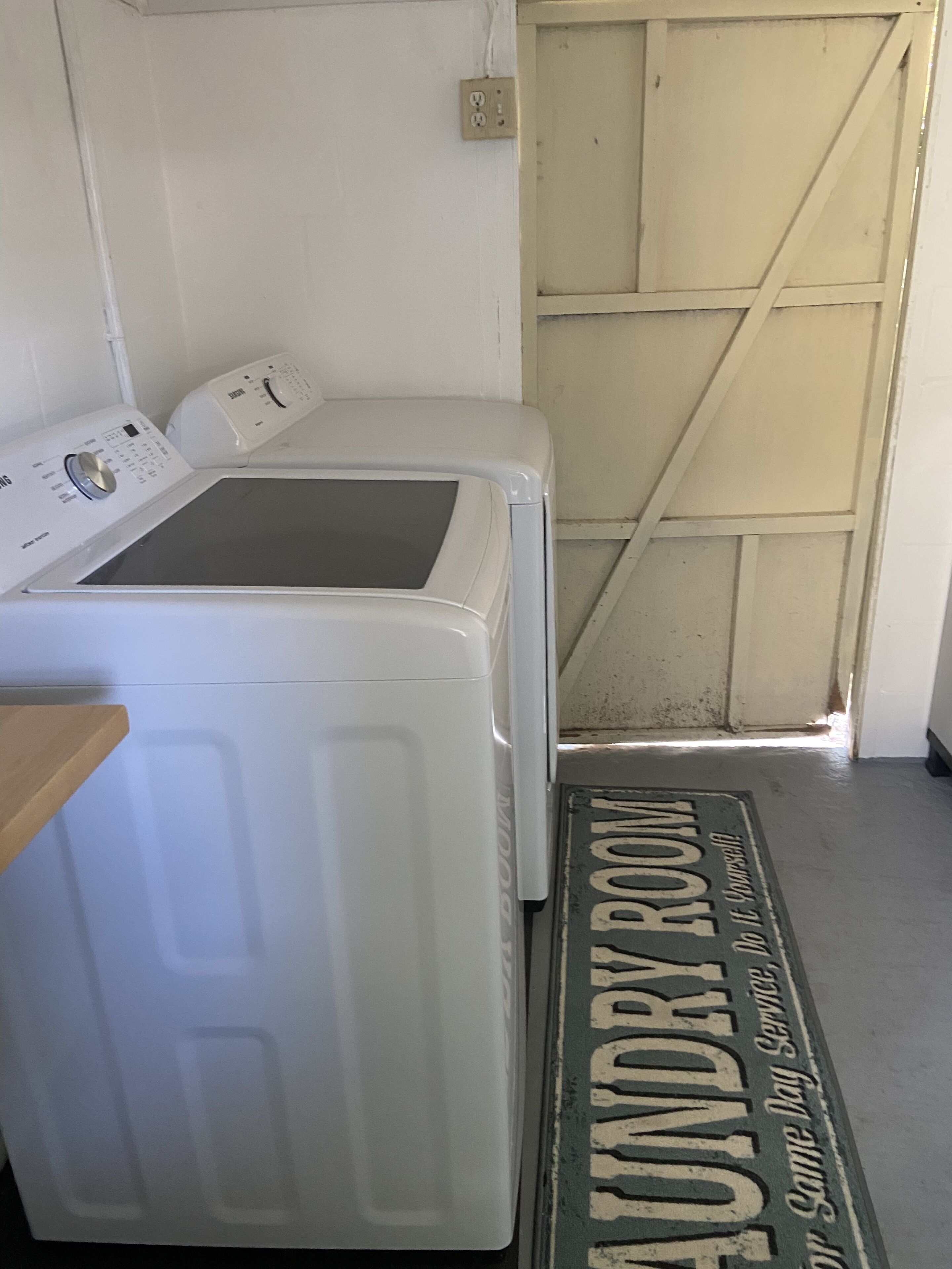 Een wasserette met een wasmachine en droger, een houten deur en een deurmat met de tekst 'Laundry Room'.