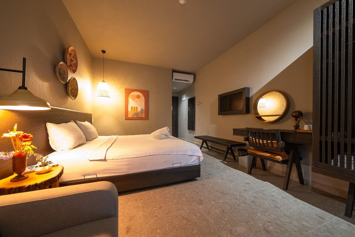 Quarto de hotel moderno com cama, mesa de cabeceira, luminária e escrivaninha com cadeira.
