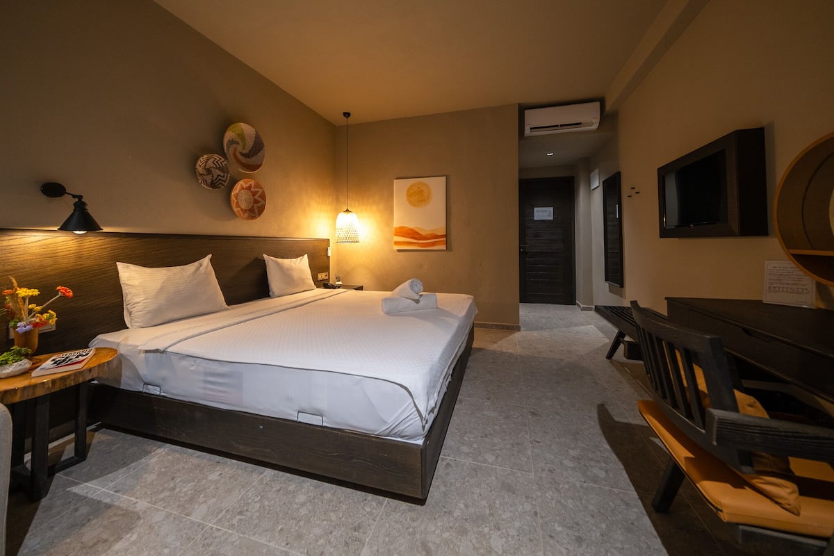 Quarto de hotel com cama, mesa de cabeceira com flores, escrivaninha e televisão.