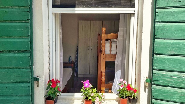 Een raam met groene luiken, een houten stoel binnenin en drie bloeiende planten in potten.
