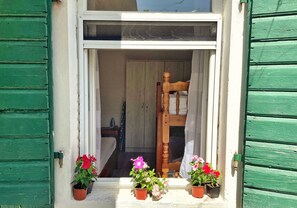 Une fenêtre avec des volets verts, une chaise en bois à l’intérieur et trois plantes en pot ornées de fleurs.