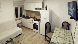 Een kleine keuken met witte kastjes, een fornuis en een koelkast. Er staat een eettafel met stoelen en een kleine potplant op het aanrecht.