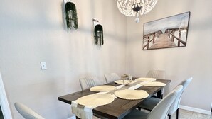 Un comedor con una mesa rectangular, seis sillas, una araña y un cuadro en la pared.