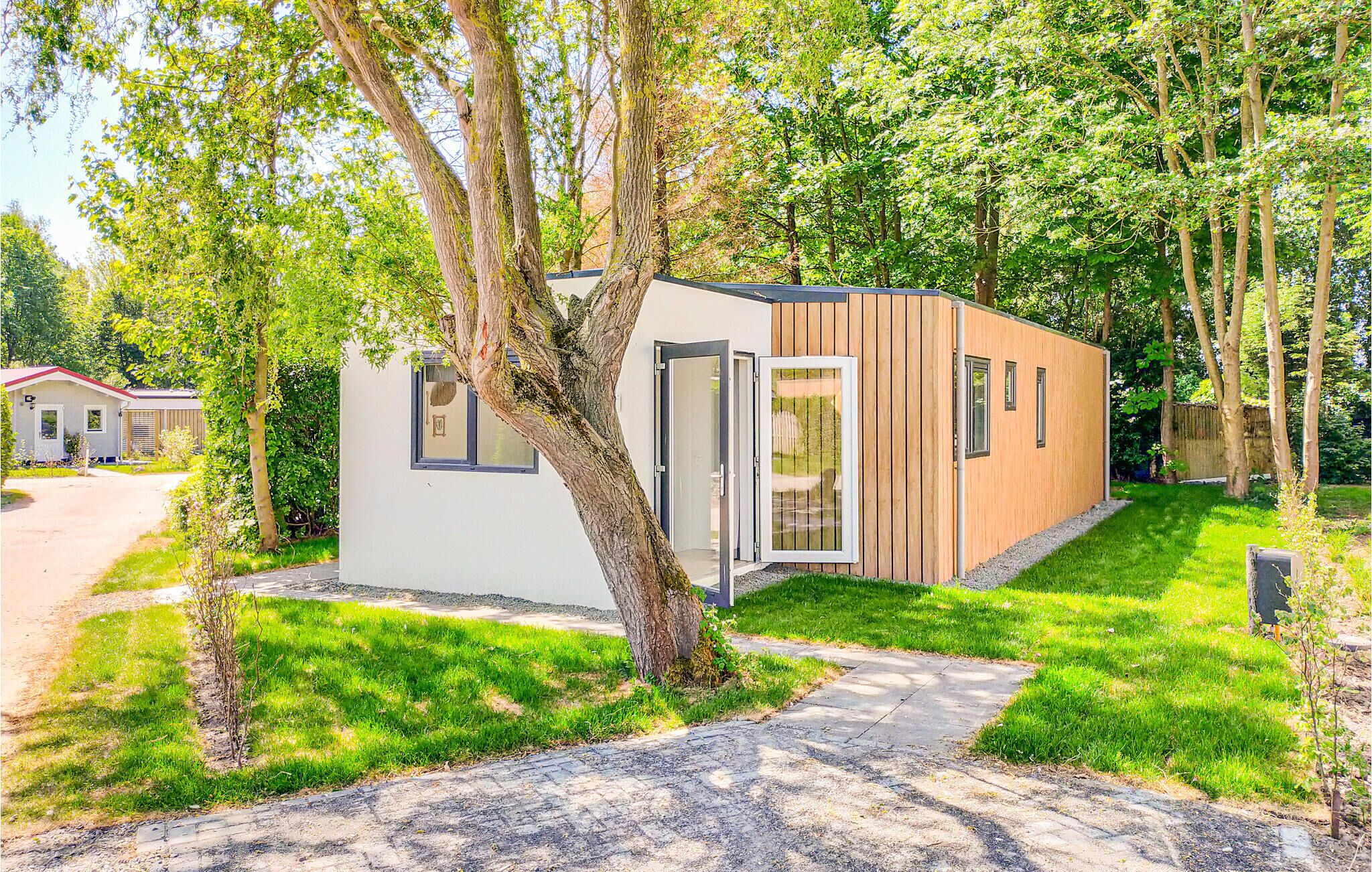 A modern, single-story cabin with a white exterior and wooden accents, surrounded by lush greenery and trees.