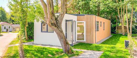 A modern, single-story cabin with a white exterior and wooden accents, surrounded by lush greenery and trees.