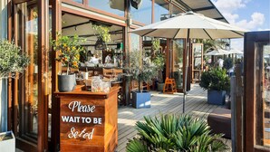 An outdoor seating area with wooden tables and chairs, a sign that reads "Please Wait to Be Set," and potted plants.