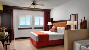 Een hotelkamer met een bed, twee bedlampjes, een plafondventilator, een bureau met een lamp, en uitzicht op de zee door grote ramen.