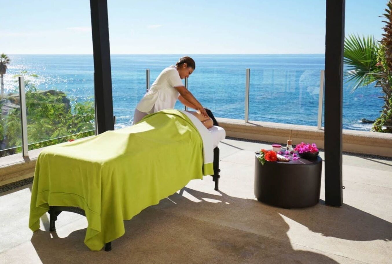 A massage table with a green cover, a massage chair, and a small table with flowers.