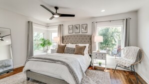 Un dormitorio con cama, mesitas de noche, una silla, ventilador de techo y ventanas amplias.